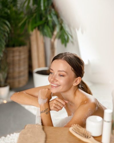 Joyful young woman smiling away and leaning on bathtub side while bathing at spa resort